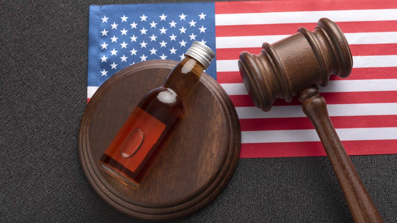 A small bottle of liquid and a judge’s gavel rest on a wooden sound block, with a United States flag in the background, symbolizing the impact of state alcohol laws on legal proceedings.