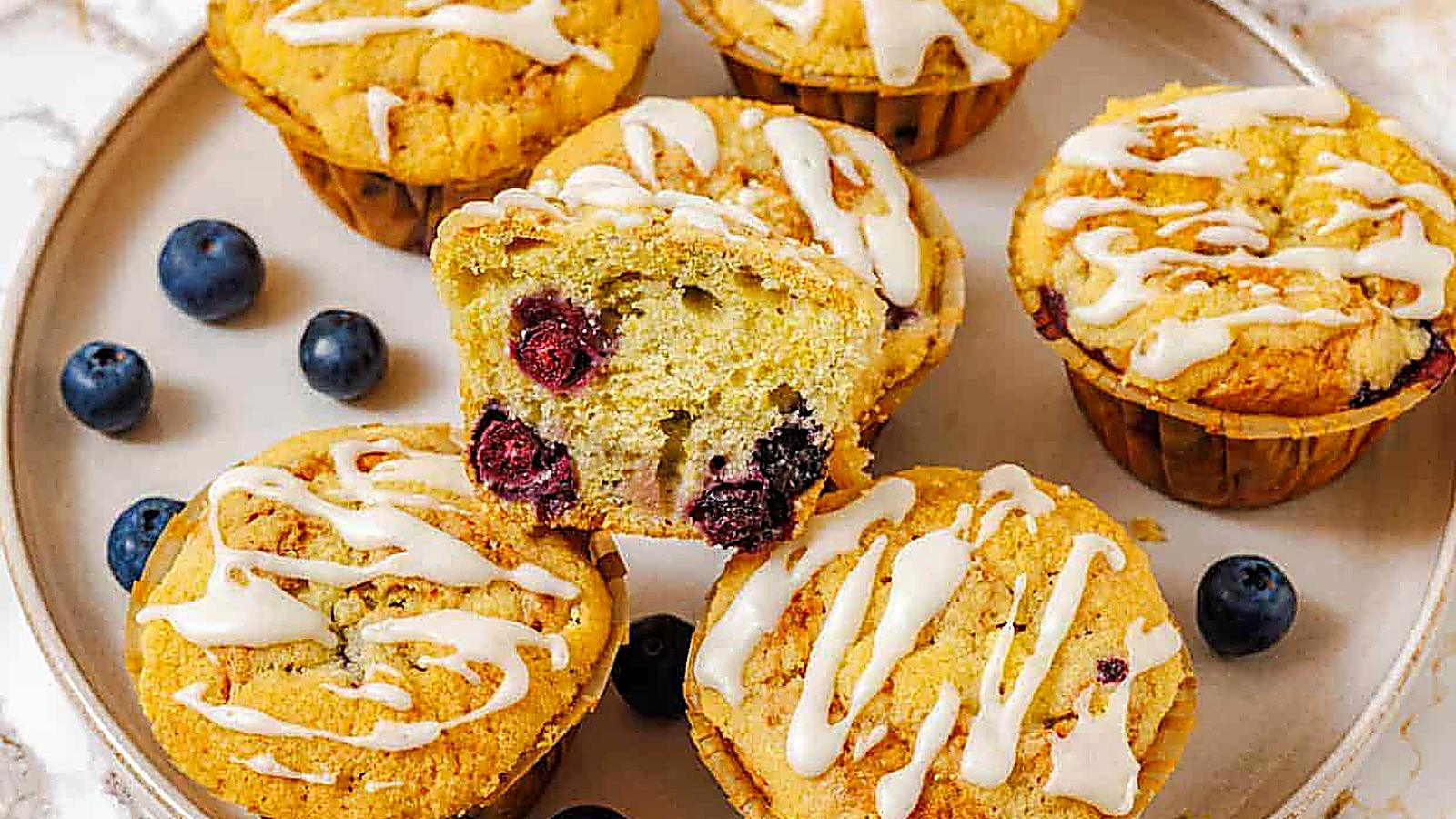A plate with five Lemon Blueberry Muffins, one muffin cut in half showing the blueberries inside, and some loose blueberries around them.