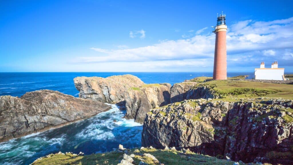 A tall lighthouse and adjacent building stand on rocky cliffs overlooking the ocean under a bright blue sky with scattered clouds.