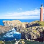 A tall lighthouse and adjacent building stand on rocky cliffs overlooking the ocean under a bright blue sky with scattered clouds.