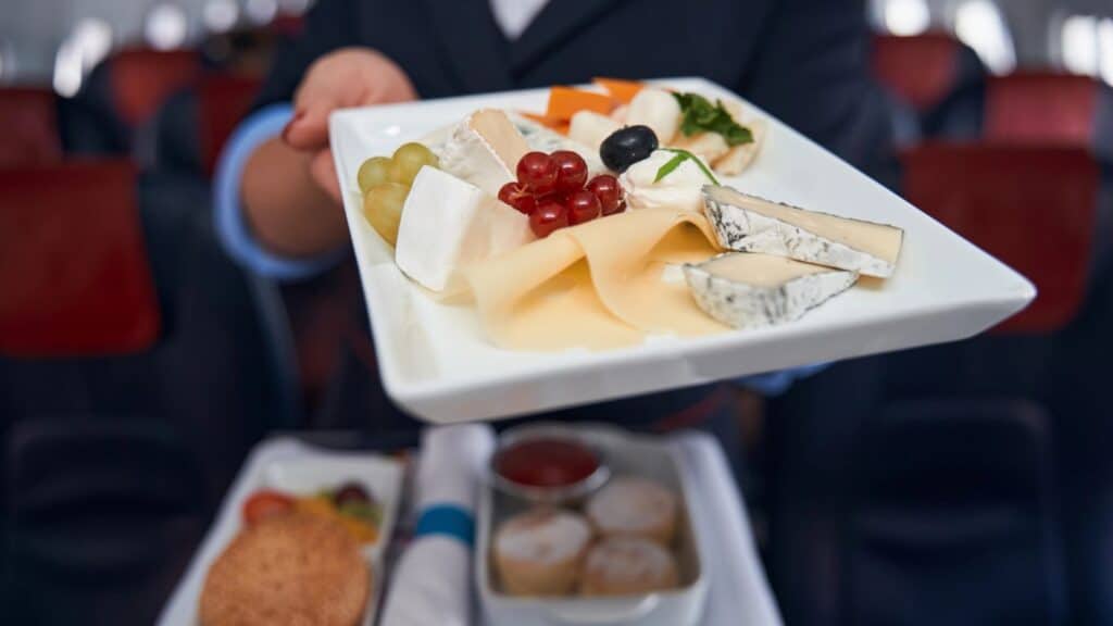 A person holds a rectangular plate with assorted cheeses, grapes, red currants, a single olive, and cheese slices on an airplane.