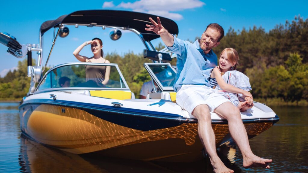 A man and girl sit on the front of a boat, smiling, with a woman standing behind them. All are enjoying a sunny day on the water with trees in the background.