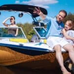 A man and girl sit on the front of a boat, smiling, with a woman standing behind them. All are enjoying a sunny day on the water with trees in the background.