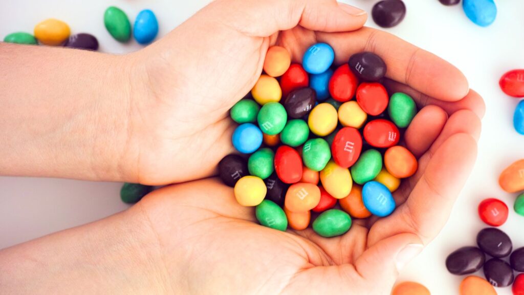 Cupped hands holding a pile of colorful candy-coated chocolates, including red, blue, green, yellow, orange, and brown pieces.