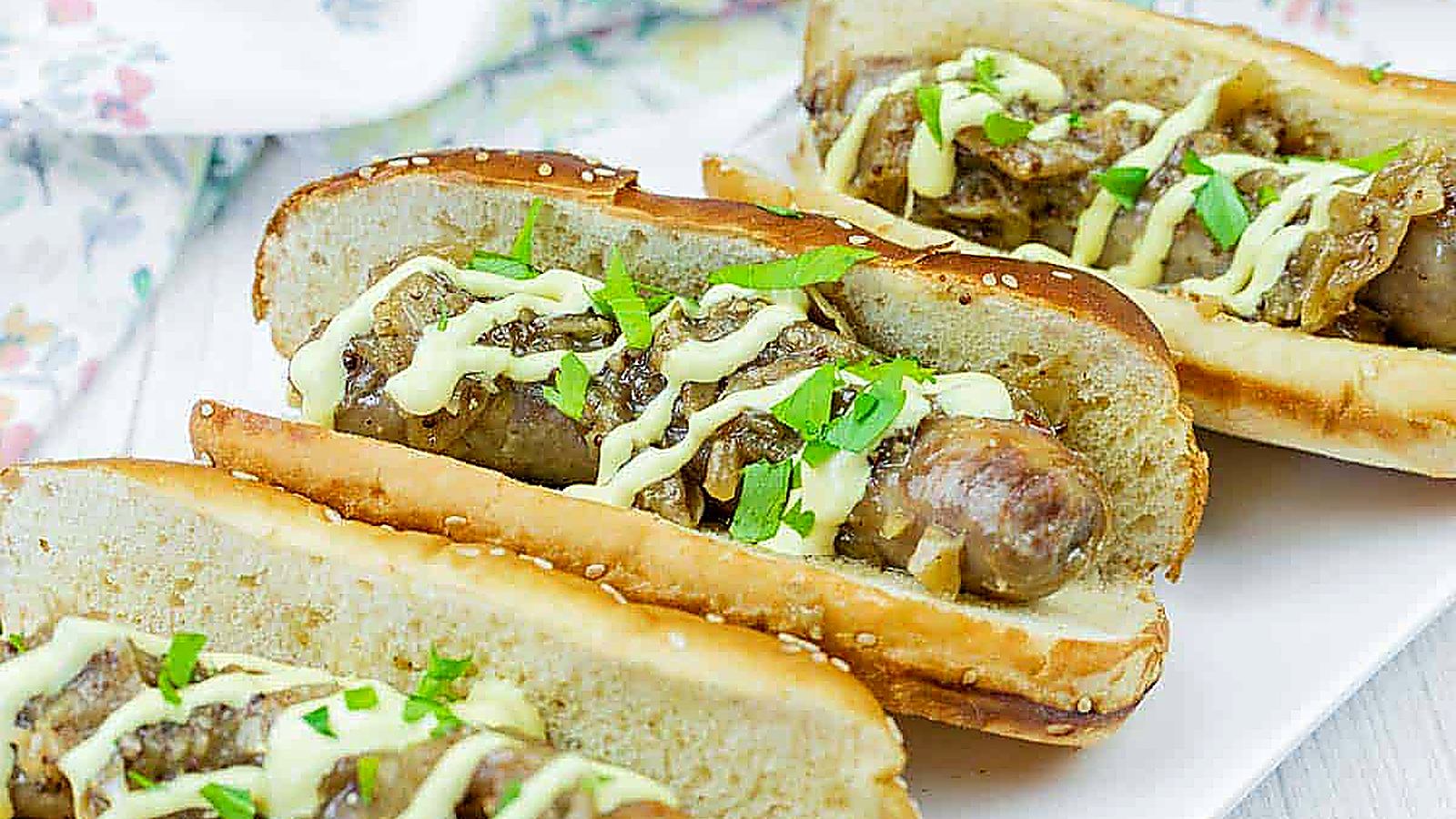 Three Beer Braised Brats in toasted buns topped with saut&eacute;ed onions, chopped parsley, and a drizzle of yellow sauce, served on a white plate.