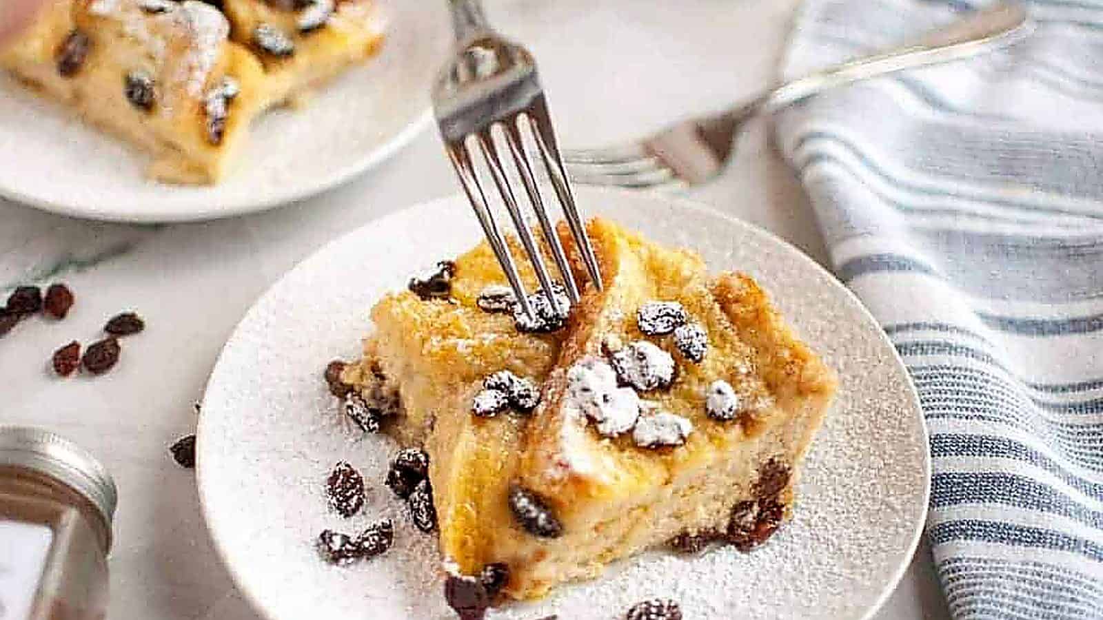 A fork cutting into a slice of bread pudding with raisins, dusted with powdered sugar, served on a white plate.