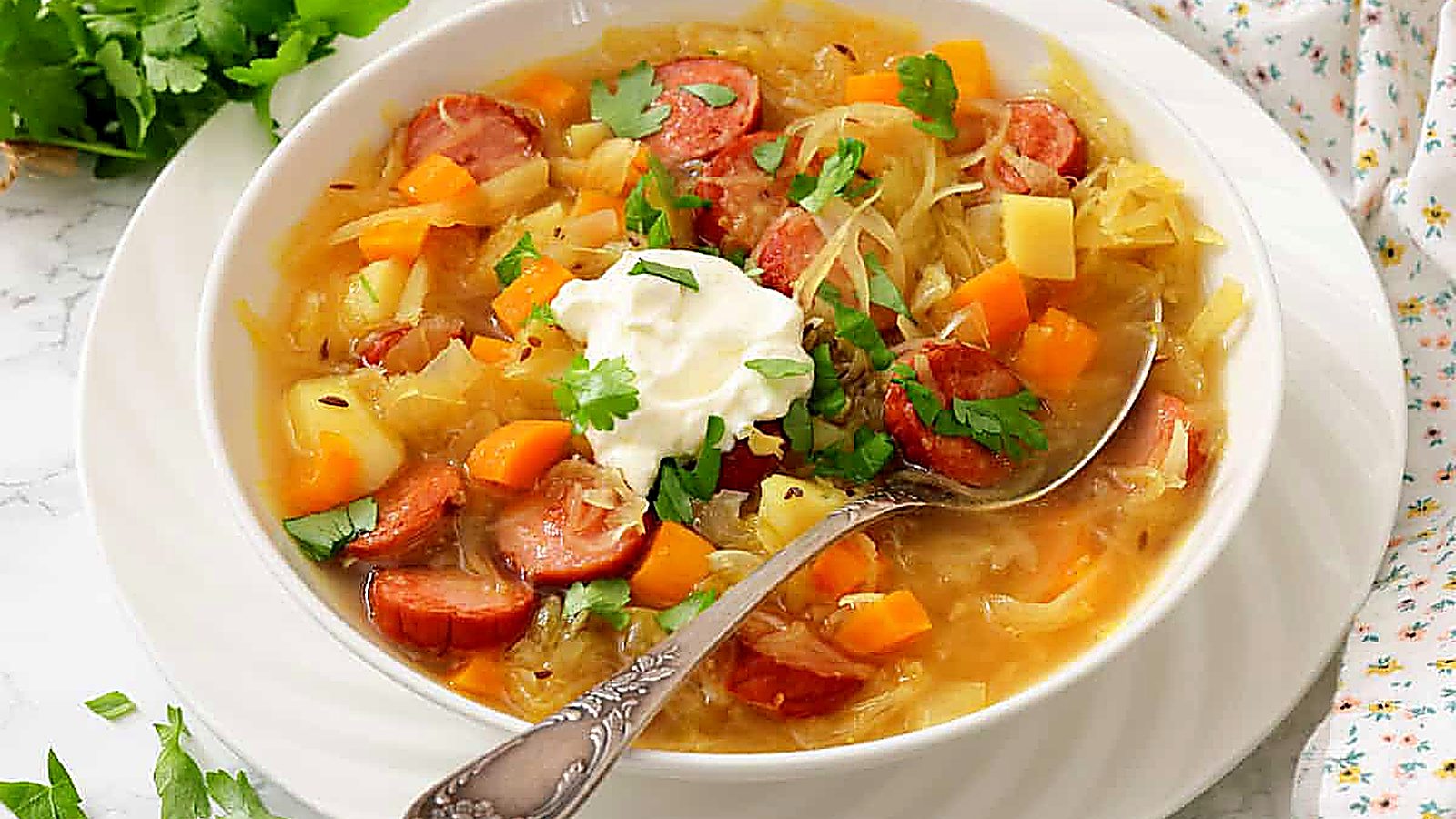 A bowl of soup with sliced sausage, potatoes, carrots, cabbage, garnished with fresh herbs and a dollop of sour cream, with a spoon resting in the bowl.