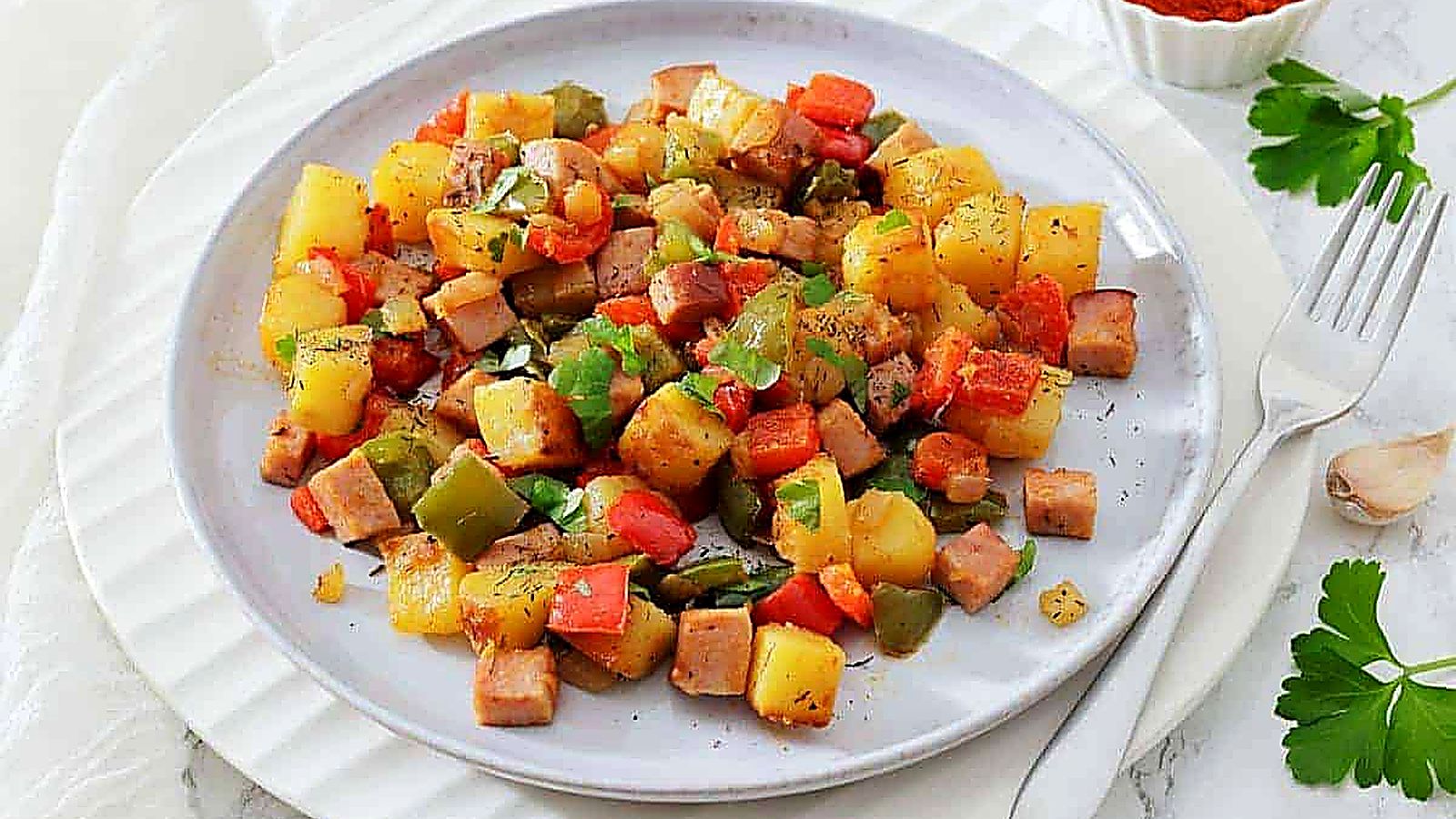 A plate of Denver Breakfast Potatoes garnished with herbs, with a fork and a dish of paprika on the side.