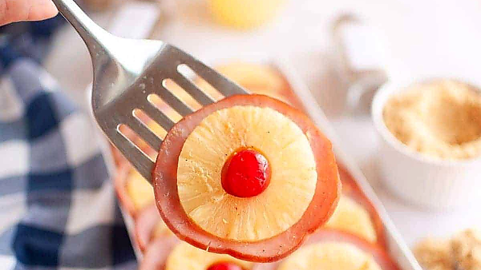 A metal spatula holds a slice of ham topped with a pineapple ring and a red maraschino cherry; more similar slices are visible in the background.