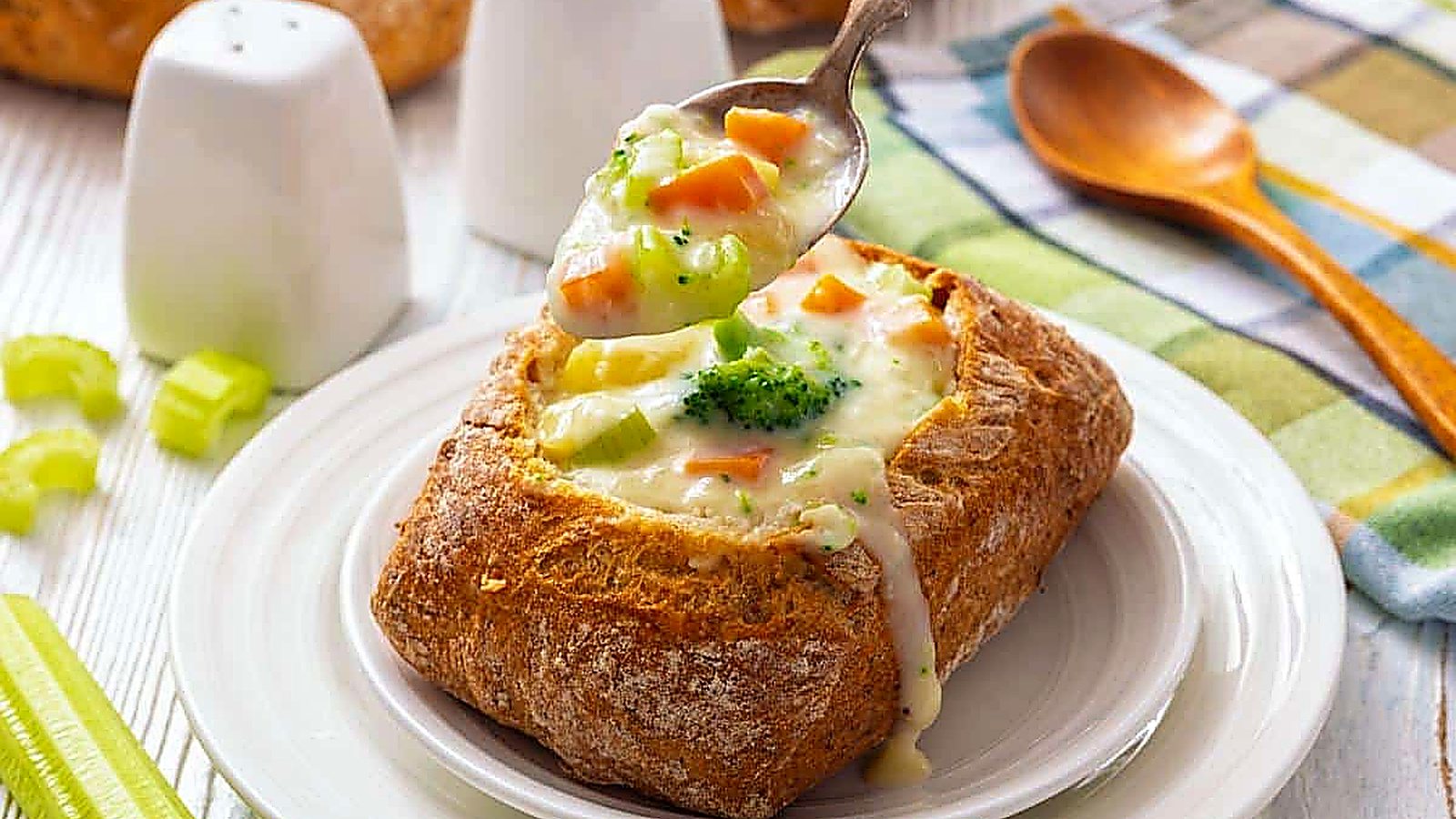 A bread bowl filled with creamy vegetable chowder, with a spoon scooping soup out; salt and pepper shakers, celery, and a wooden spoon are nearby.
