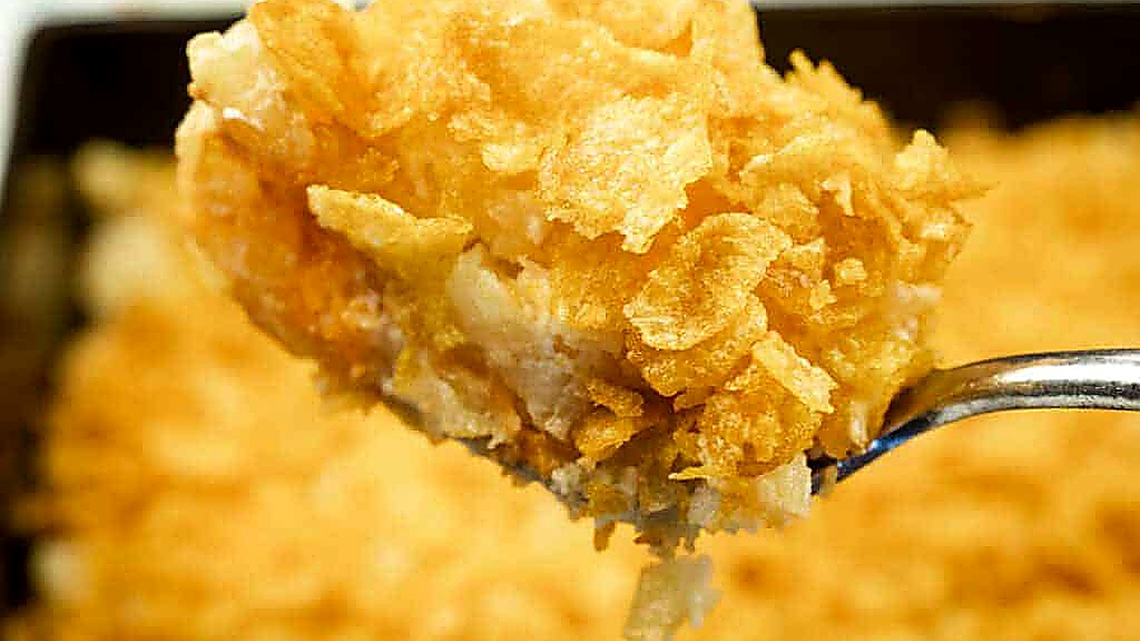 A close-up of a spoonful of Funeral potatoes with Cream Cheese, topped with crushed cornflakes, with the rest of the casserole in the background.