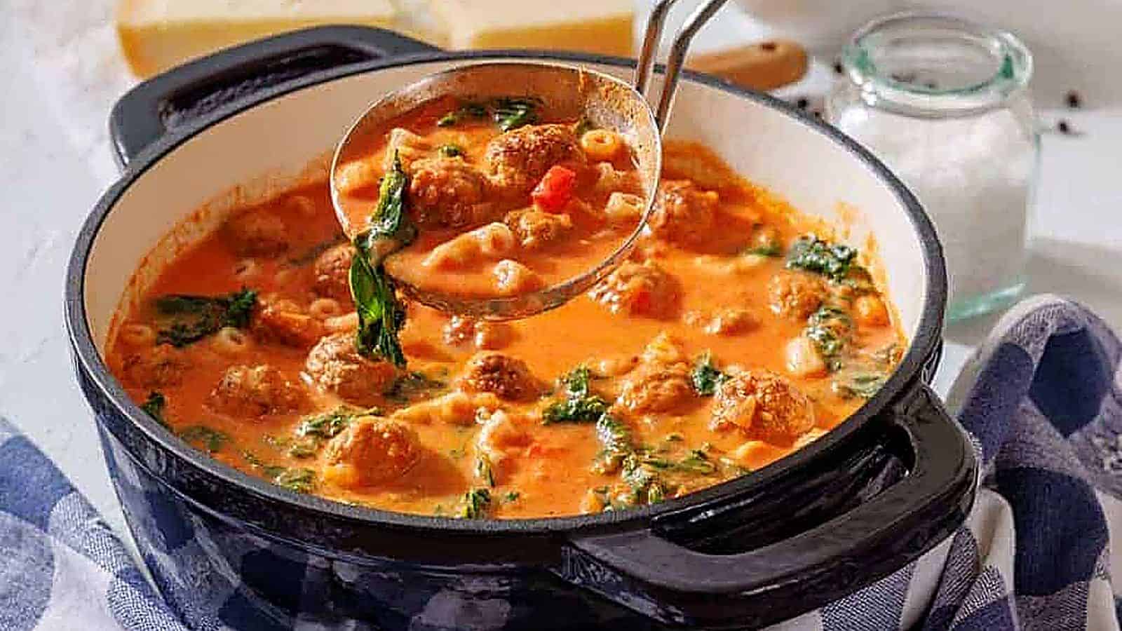 A ladle lifts meatball soup with spinach, pasta, and tomato broth from a black pot on a kitchen counter.
