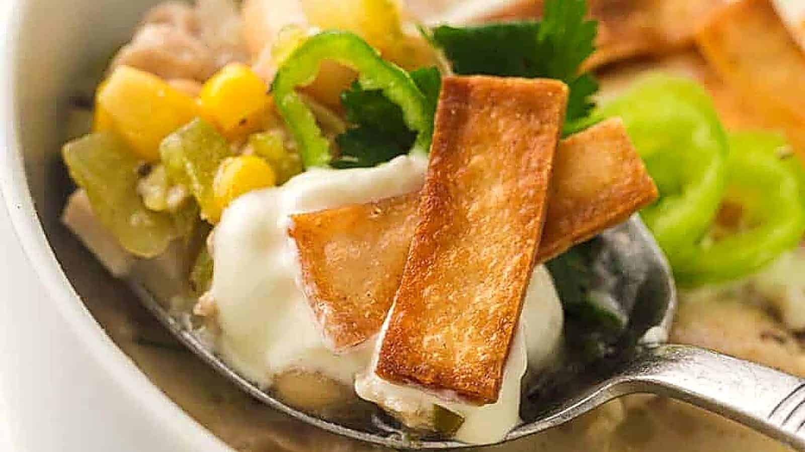 A spoonful of white chili with sour cream, corn, green chili slices, cilantro, and crispy tortilla strips.
