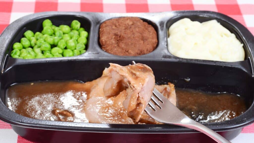 A TV dinner tray with peas, mashed potatoes, a brownie, and sliced meat with brown gravy, on a red and white checkered tablecloth.