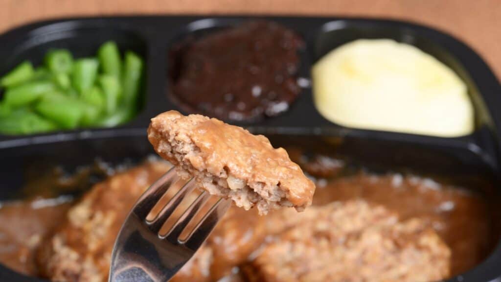 A fork holds a piece of Salisbury steak with gravy. In the divided tray are mashed potatoes, green beans, and a brown dessert.