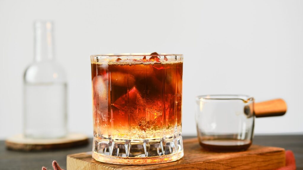 A glass filled with a dark iced espresso tonic sits on a wooden tray next to a small glass pitcher and a clear bottle in the background.