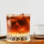 A glass filled with a dark iced espresso tonic sits on a wooden tray next to a small glass pitcher and a clear bottle in the background.