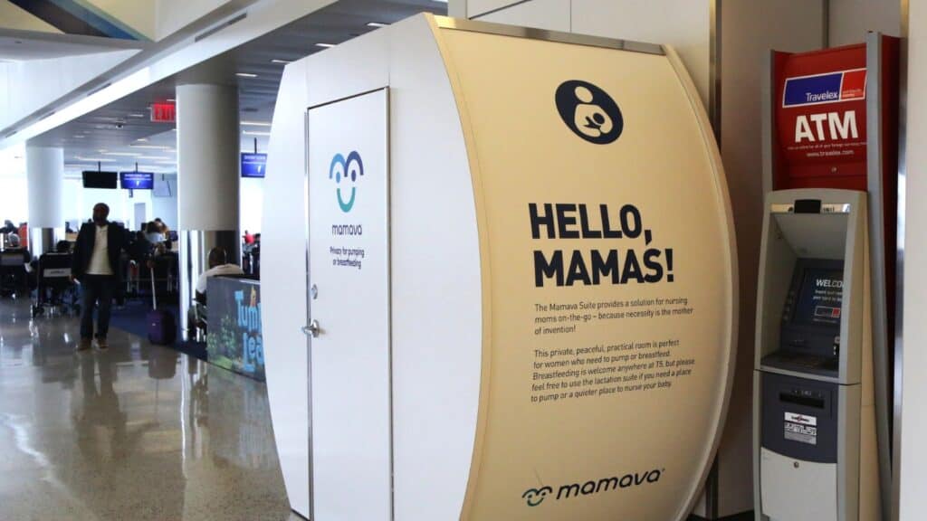 White Mamava lactation pod with &ldquo;Hello, Mamas!&rdquo; sign next to a red ATM in an airport terminal&mdash;an amenity you don&rsquo;t expect in an airport; people and gates visible in the background.
