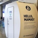White Mamava lactation pod with “Hello, Mamas!” sign next to a red ATM in an airport terminal—an amenity you don’t expect in an airport; people and gates visible in the background.