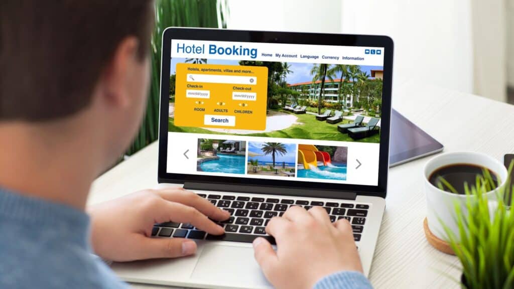 Person using a laptop to search for hotels on a booking website; room search fields, hotel images, and helpful US hotel fee facts are visible on the screen.