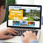 Person using a laptop to search for hotels on a booking website; room search fields, hotel images, and helpful US hotel fee facts are visible on the screen.