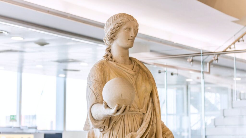 A marble statue of a robed woman holding a sphere stands in a modern, glass-walled interior space with natural light&mdash;an elegant presence you don&rsquo;t expect in an airport.