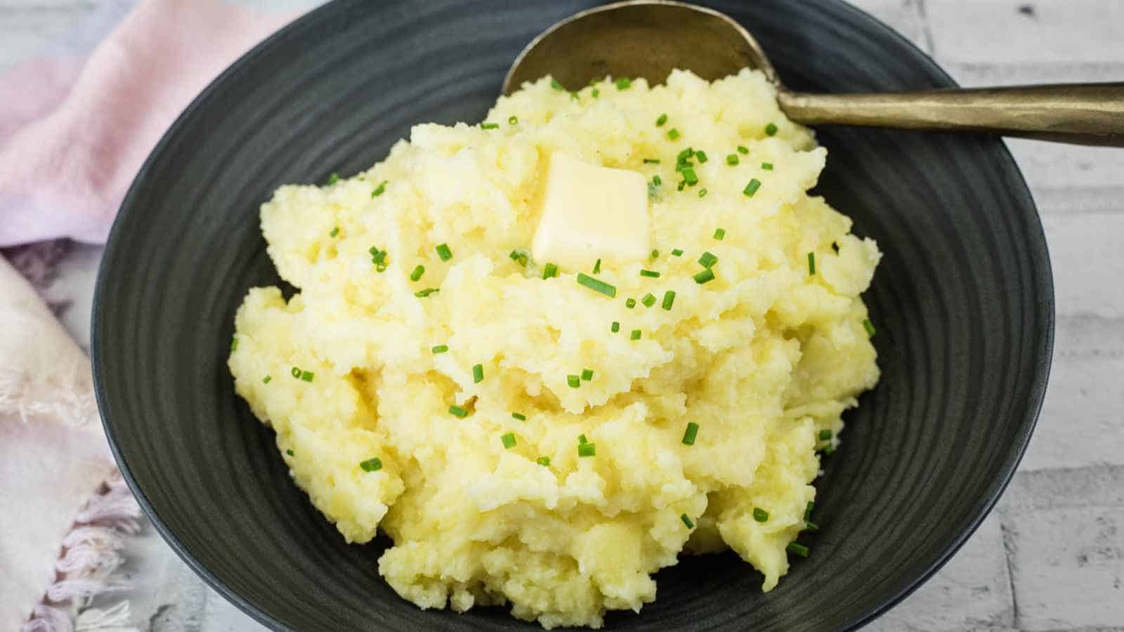 A bowl of mashed potatoes topped with a pat of butter and chopped chives, with a spoon resting on the side of the bowl.