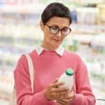 A woman wearing glasses and a pink sweater stands in a grocery store, reading the label on a bottle in her hand, with shelves of dairy products blurred in the background.