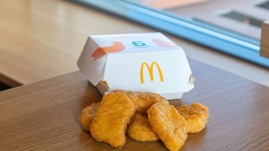 Six McDonald&rsquo;s chicken nuggets are displayed on a wooden table with their open paper box in the background near a window.