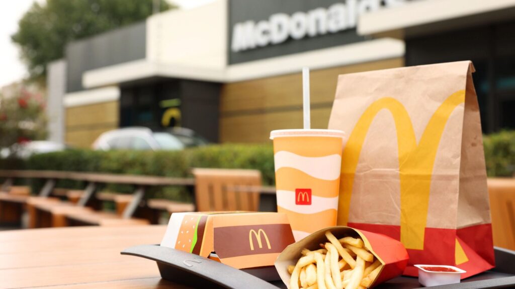 McDonald&rsquo;s meal on an outdoor table, featuring fries, a drink, a burger box, and a paper bag with the McDonald&rsquo;s logo, with a McDonald&rsquo;s restaurant in the background.