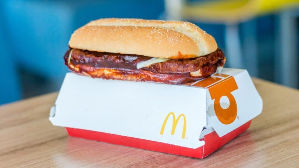 A McDonald&rsquo;s McRib sandwich with barbecue sauce and onions sits on top of its branded box on a wooden table&mdash;a tasty example of how some fast-food items started locally before gaining nationwide popularity.