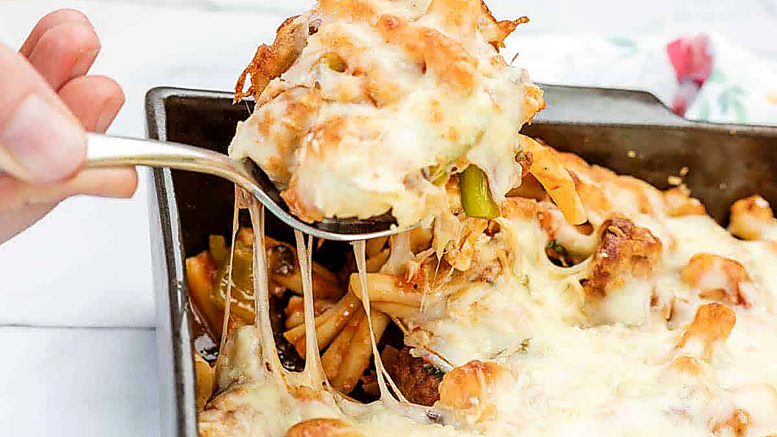 A hand lifts a serving of meatball casserole from a rectangular baking dish, showing melted cheese stretching.