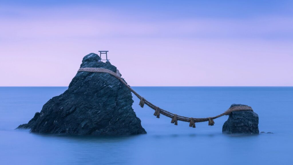Two rocky sea stacks connected by a thick rope with tassels, with a small torii gate on the larger rock, under a blue sky at dawn—a beautiful reminder that Japan is more than Kyoto and Tokyo.