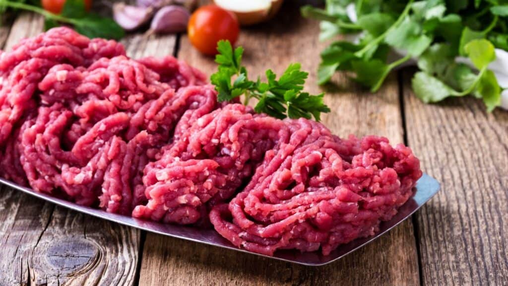 Raw ground beef&mdash;one of several grocery items whose prices rose&mdash;rests on a metal tray, garnished with parsley, atop a wooden table surrounded by fresh vegetables and herbs in the background.