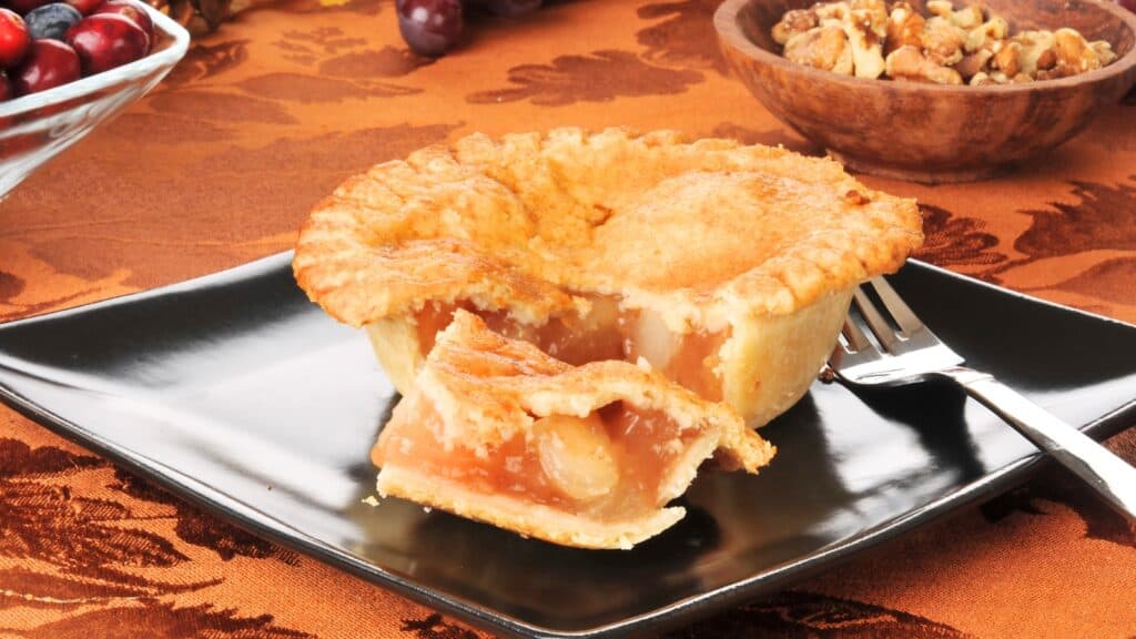 A slice of apple pie with a flaky crust sits on a black plate with a fork, with nuts and fruit in the background on an orange tablecloth—a tempting treat for those baking less at home but still craving homemade flavor.