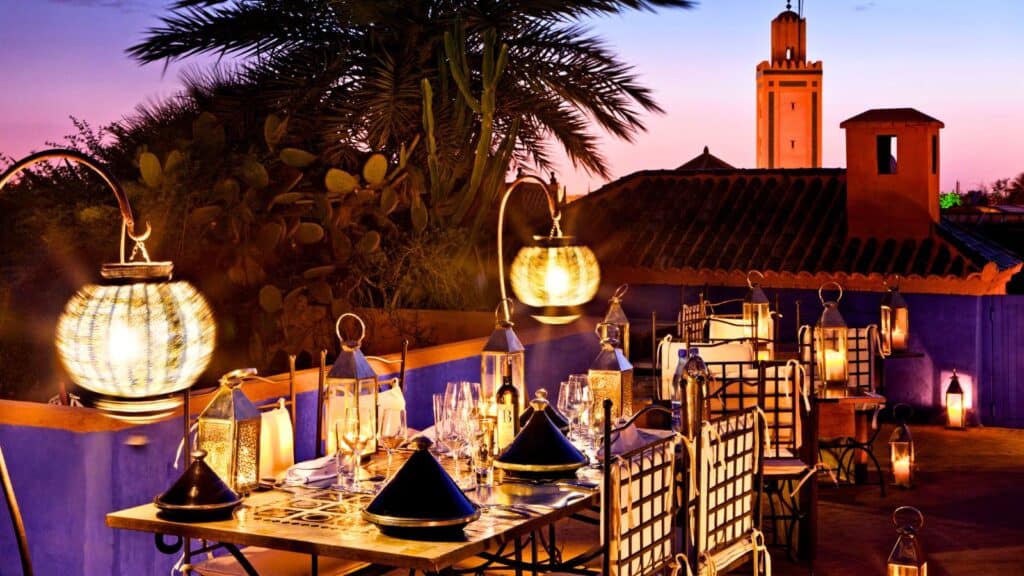 A rooftop restaurant at sunset with lantern-lit tables, traditional Moroccan decor, palm trees, and a tower visible in the background.