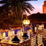 A rooftop restaurant at sunset with lantern-lit tables, traditional Moroccan decor, palm trees, and a tower visible in the background.