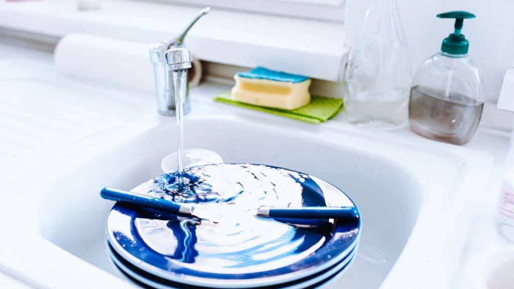 Blue and white plates with cutlery are stacked in a white kitchen sink under running water. A sponge, soap dispenser, and towel are on the counter nearby.