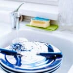 Blue and white plates with cutlery are stacked in a white kitchen sink under running water. A sponge, soap dispenser, and towel are on the counter nearby.