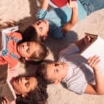 Four children lie on a light carpet in a circle, each holding and reading a book from Dolly Parton&rsquo;s Imagination Library, with sunlight shining on them.