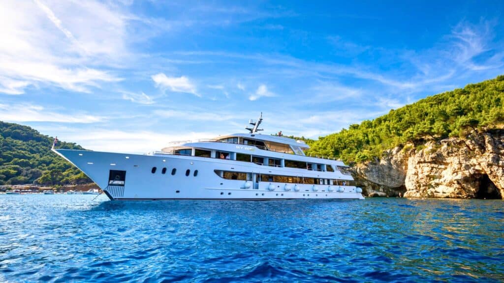 A large white yacht is anchored on blue water near a rocky, tree-covered coastline under a partly cloudy sky.