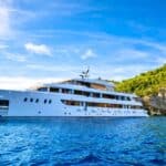 A large white yacht is anchored on blue water near a rocky, tree-covered coastline under a partly cloudy sky.