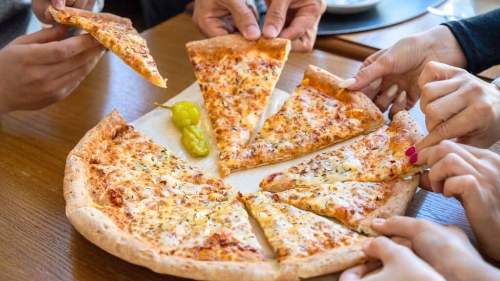 Several people take slices of cheese pizza from a shared pizza on a wooden table, with two yellow peppers on the pizza—celebrating National Cheese Pizza Day together.