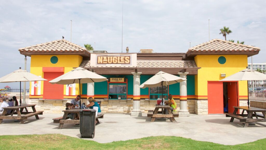 Colorful fast-food restaurant with “Naugles” sign, outdoor seating, and umbrellas. Once a staple among restaurant chains that faded away, it now hosts people sitting at picnic tables on a sunny day.