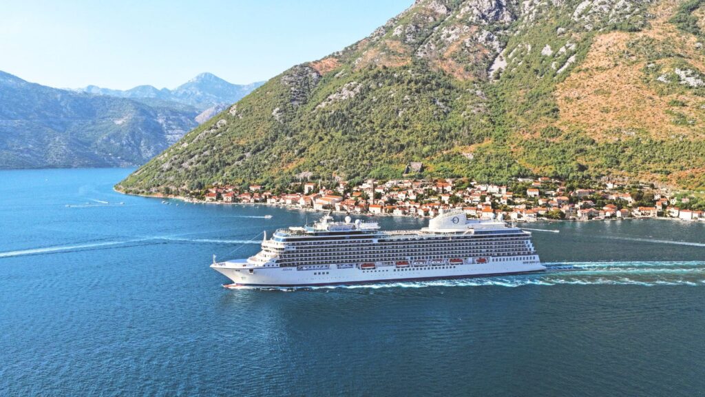 A white cruise ship sails through a narrow blue waterway, passing a small coastal village with houses at the base of green, rocky mountains.