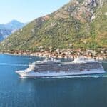 A white cruise ship sails through a narrow blue waterway, passing a small coastal village with houses at the base of green, rocky mountains.