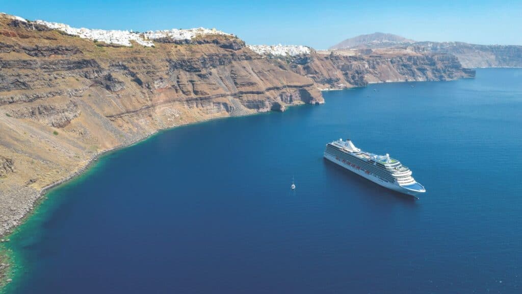 A cruise ship is anchored in blue water near steep coastal cliffs with white buildings on top under a clear sky.