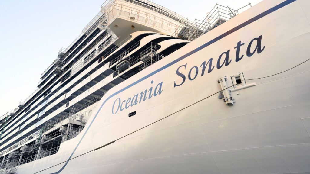 Large white cruise ship named “Oceania Sonata” docked at port, viewed from a low angle. The ship’s name is visible on the hull, with multiple balconies and exterior scaffolding.
