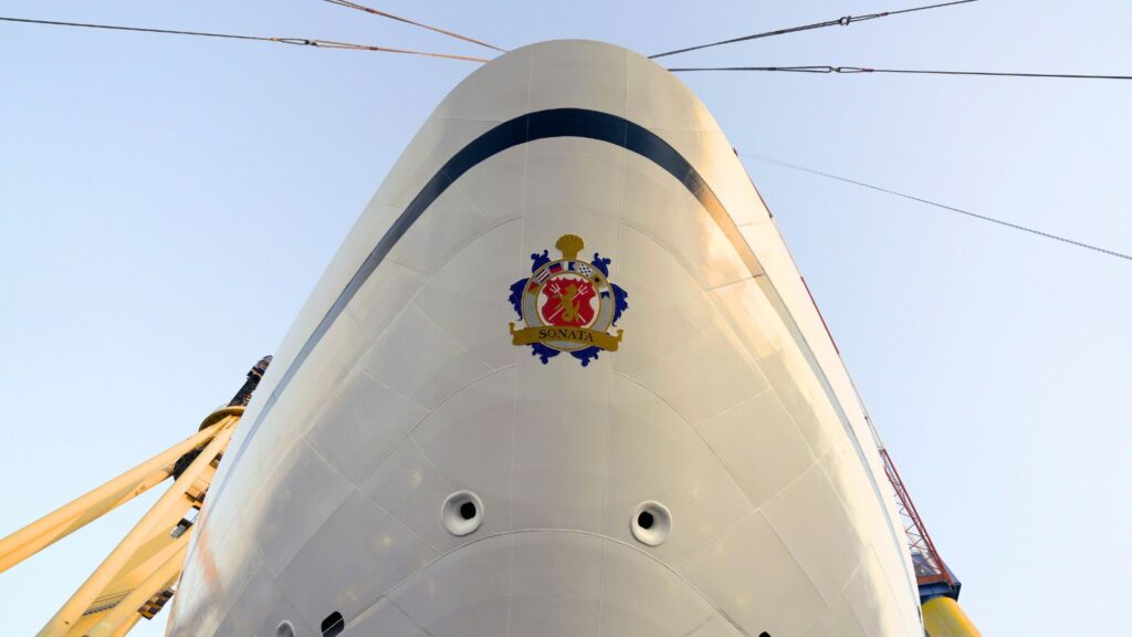 Close-up view of the bow of a white ship with a crest labeled “SONATA,” cables extending from the sides, and clear sky in the background.