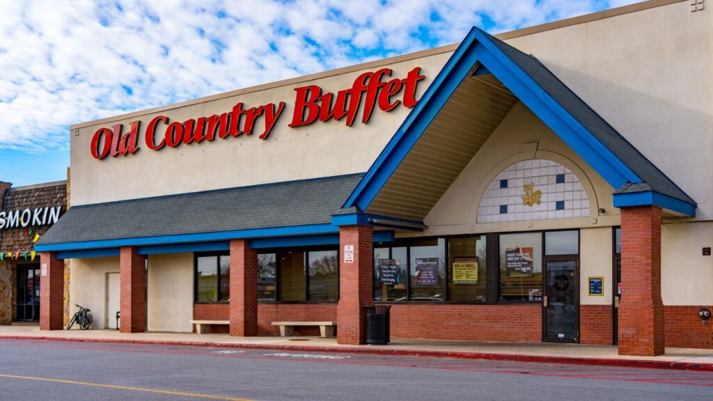 Exterior of an Old Country Buffet restaurant, once one of the well-known salad bar chains, with red signage, blue roof accents, brick walls, large windows, and a mostly empty parking area in front.